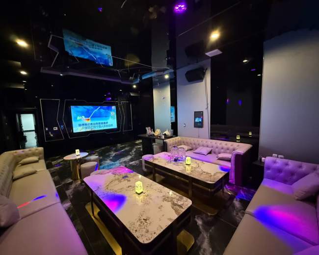 The image shows a modern lounge area with dark decor, featuring large sofas and tables, illuminated by colorful lights and a large screen on the wall.