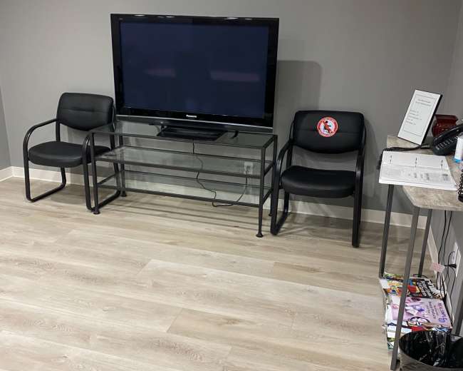 A waiting area with two black chairs beside a glass TV stand, which holds a television against a gray wall, with a magazine rack and a trash bin nearby.