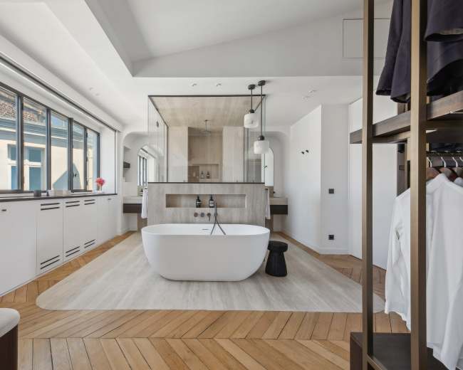 The image shows a modern bathroom featuring a freestanding white bathtub, a wooden floor, and large windows that allow natural light to enter the space.
