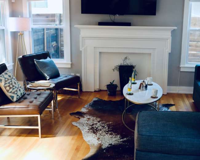 The image shows a cozy living room with a blue couch, a leather chair, a coffee table, and a decorative rug on a wooden floor, illuminated by natural light from windows.