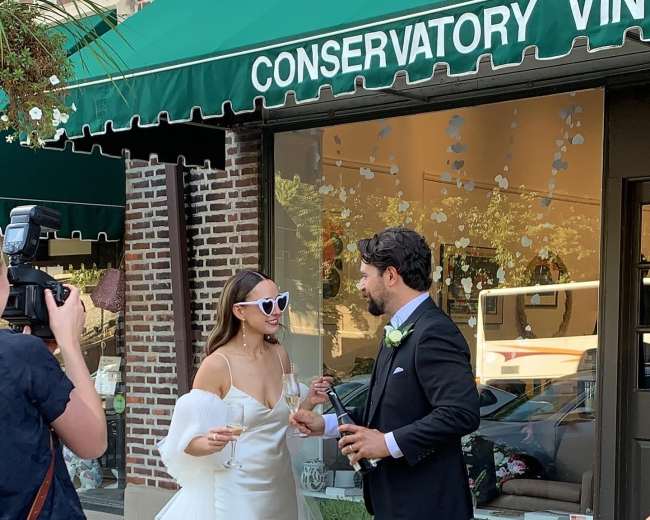 A bride in a sleek wedding dress and oversized sunglasses shares a toast with a groom in a suit outside a storefront.