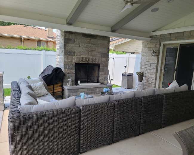 A covered patio area featuring a large sectional couch arranged around a stone fireplace.