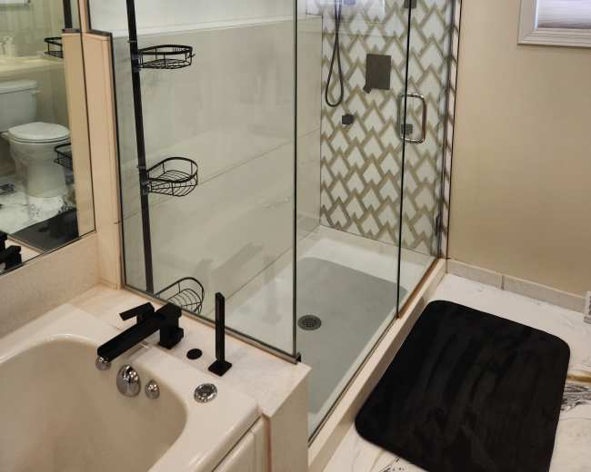 The image shows a modern bathroom with a glass shower enclosure featuring a patterned tile background, a freestanding bathtub, and shelving for toiletries.