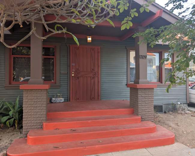 The image shows a home's entrance with a red door, flanked by two pillars and steps leading up to it, surrounded by greenery.