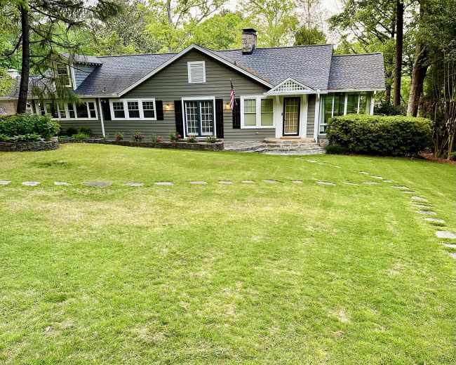 A gray, two-story house with white trim is set amid a lush green lawn, featuring a stone pathway leading to the entrance.