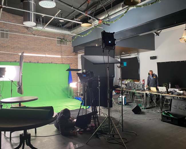 A studio space is set up with a green screen, lighting equipment, and several workstations, while a person is working on a laptop on the floor.