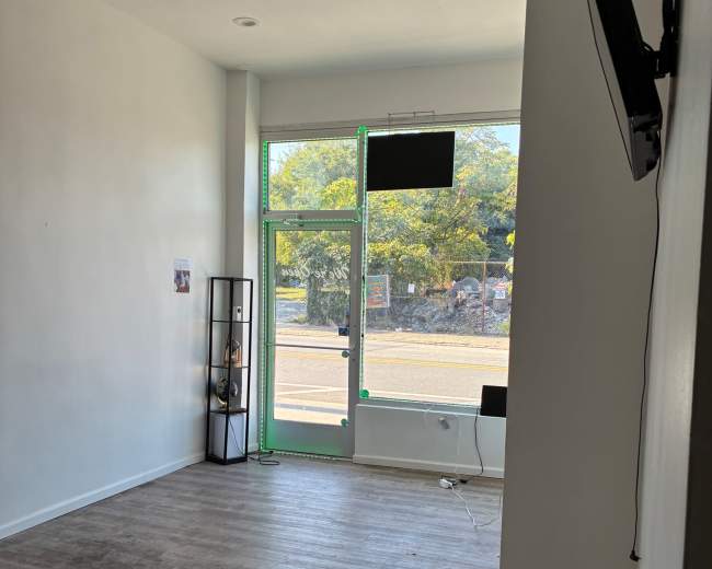 The image shows an empty, light-filled interior space with a window facing a street, a television mounted on the wall, and minimal furniture.