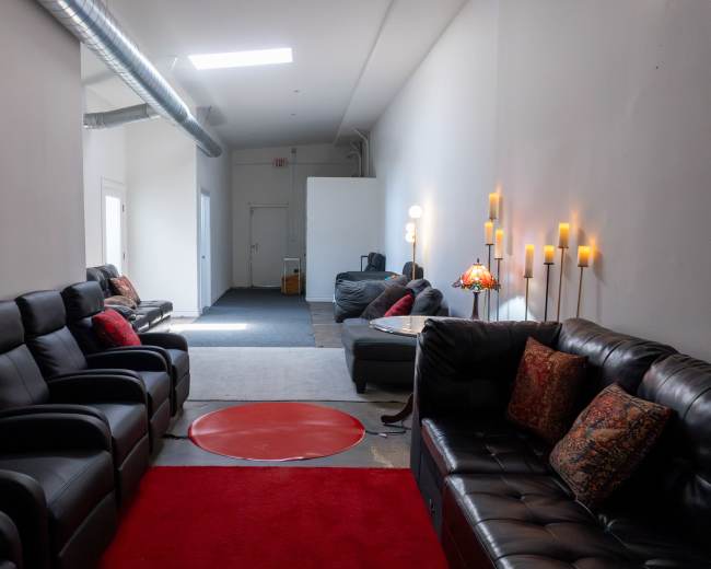 The image shows a long hallway featuring black leather sofas on either side, a red circular rug in the center, and soft lighting from wall-mounted candles.
