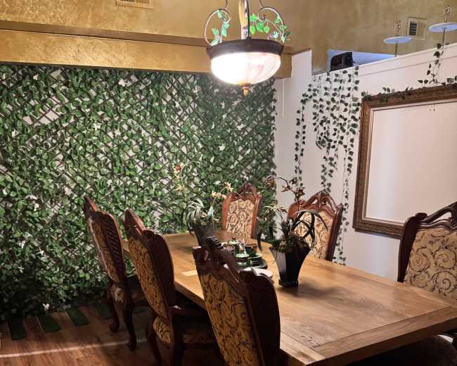 The image shows a dining area with a large wooden table surrounded by ornate chairs, set against a background of greenery and a decorative mirror.