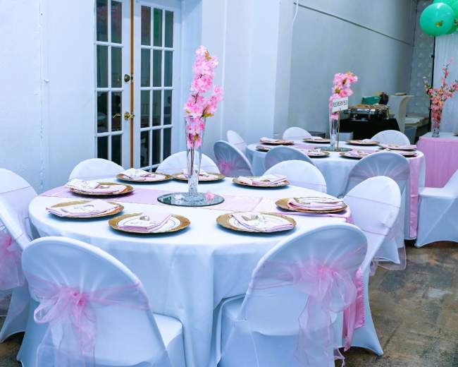 A banquet setup with round tables covered in white tablecloths, each adorned with pink decorations and centerpieces featuring floral arrangements.