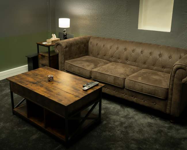 A brown tufted sofa and a wooden coffee table are arranged on a carpeted floor in a dimly lit room with muted green and gray walls.