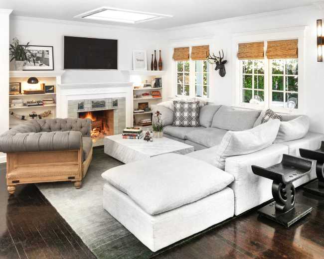 The image shows a cozy living room featuring a large gray sectional sofa, a fireplace with a TV above it, and bookshelves filled with decorative items.