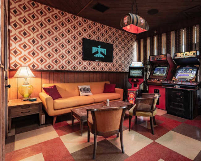 The image shows a retro-style game room featuring a brown sofa, two chairs, a wooden table, and several vintage arcade machines against a patterned wall.