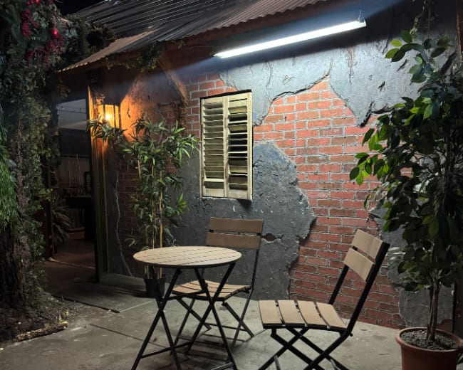 The image shows a small outdoor seating area with two folding chairs and a round table beside a rustic-looking wall featuring bricks and a window with shutters.