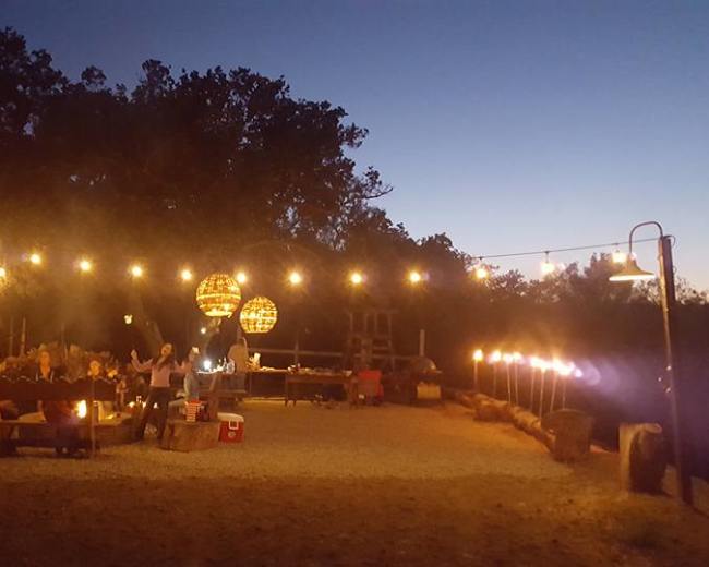 A camping area is illuminated by string lights and lanterns, featuring a seating arrangement around a fire pit.