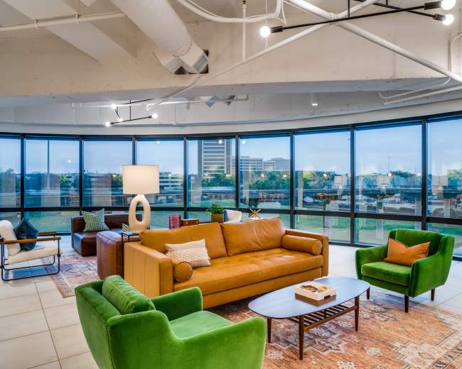 The image shows a modern, open lounge area with large windows, featuring a yellow sofa, green armchairs, a coffee table, and a distinctive lamp, set against an urban backdrop.