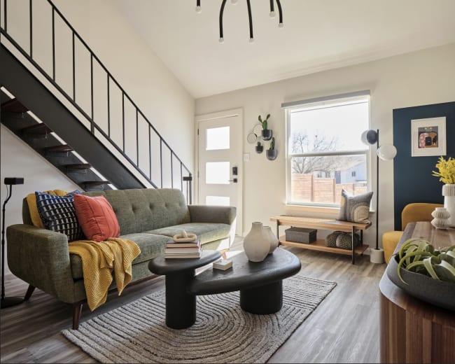 The living room features a green sofa, a round black coffee table, and a staircase, with large windows letting in natural light.