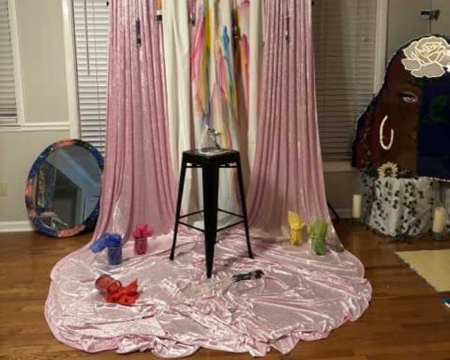 The scene features a pink draped backdrop with colorful streamers, a black stool at the center, and various art supplies scattered on the floor.