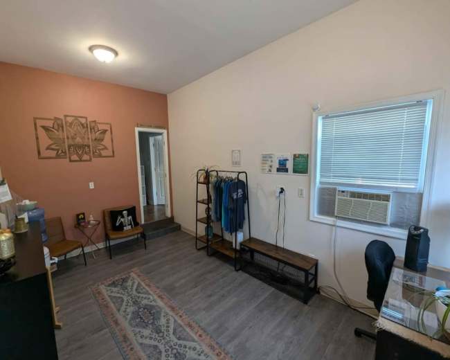 The image shows a neatly organized room with a small desk, a shelf, an air conditioning unit, and decorative wall art.
