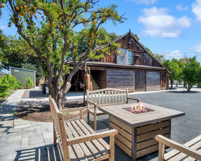 A rustic wooden building serves as a backdrop for a patio area featuring a fire pit and benches, surrounded by trees and landscaped greenery.