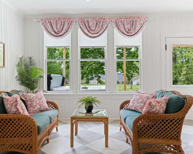 A cozy seating area features two wicker sofas with floral cushions, a glass coffee table, and large windows framed by patterned curtains, allowing natural light to fill the space.