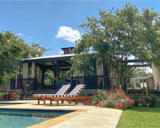 A swimming pool with lounge chairs is featured in a backyard surrounded by trees and flowers, with a covered patio area in the background.