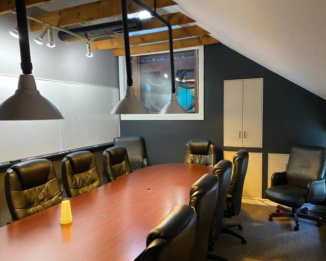 The image shows a conference room with a long wooden table, black leather chairs, and pendant lights hanging from a sloped ceiling.