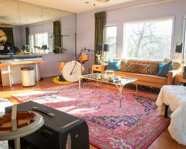 The image shows a cozy living room with a golden sofa, a glass coffee table on a patterned rug, a piano, and musical instruments visible near large windows.