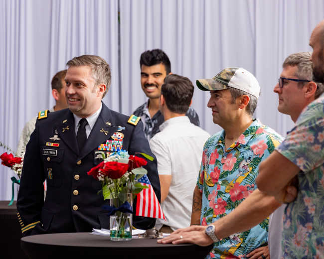 A group of men in a social setting engages in conversation, with one man in a military uniform and others in casual attire, surrounded by decorative flowers and American flags.