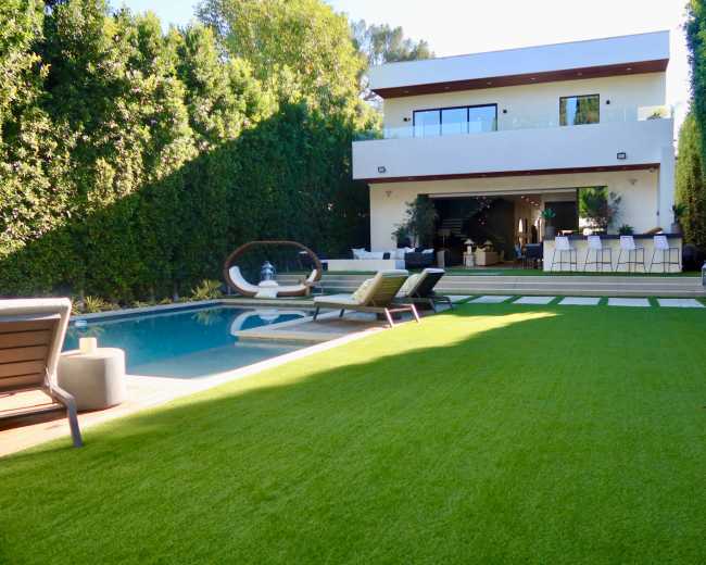 A modern two-story house with large windows overlooks a neatly landscaped backyard featuring a swimming pool and lounging area.