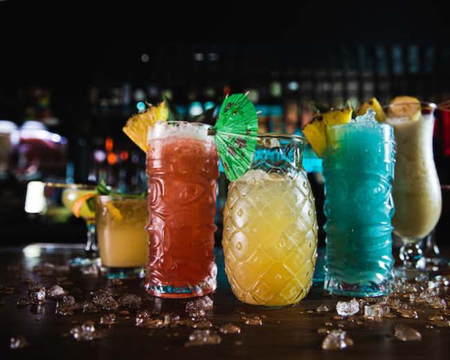 A variety of colorful cocktails are displayed on a bar counter, surrounded by scattered ice and garnished with fruit and umbrellas.
