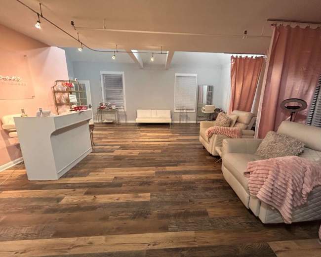 The image shows a modern lounge area with light wooden flooring, two white leather recliners, and a small reception bar in a softly lit room decorated with pink curtains and accents.