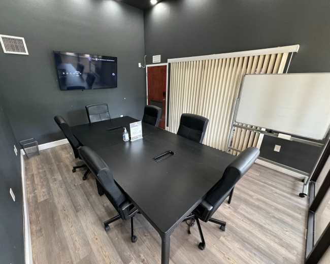A conference room features a large black table surrounded by black chairs, with a whiteboard and a television mounted on the wall.