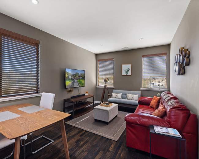 The image shows a modern living room with a red sofa, a wooden dining table, and a television mounted on the wall, along with large windows featuring wooden blinds.