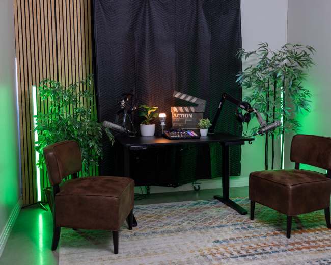 A well-lit recording studio setup features two brown chairs facing a desk with a film clapperboard and potted plant, surrounded by green-lit plants.