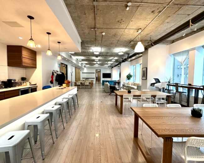 The image shows an open-concept modern office space with a long bar, seating areas, and large windows.