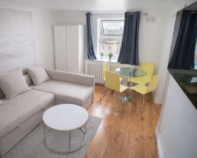 The image shows a small living area with a grey sofa, a round white coffee table, and a glass dining table with four yellow chairs, complemented by a window and a wall featuring a picture of a bridge.