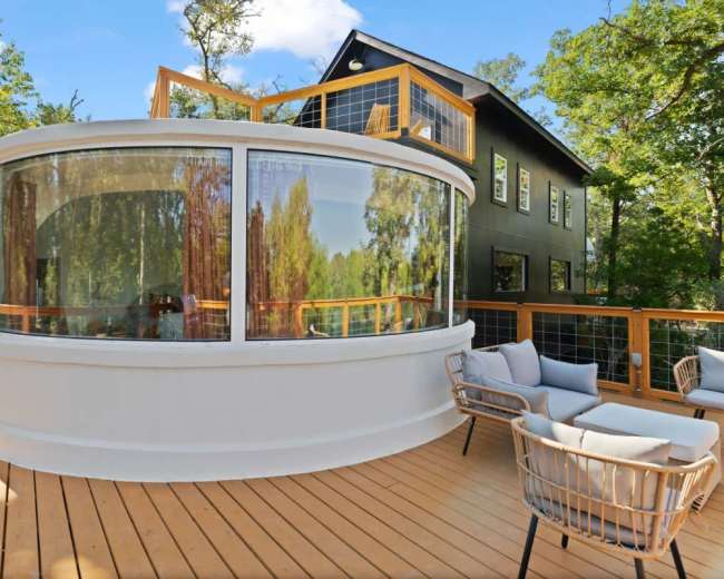 The image shows a modern outdoor deck featuring a round, glass-enclosed seating area and a contemporary building surrounded by trees.