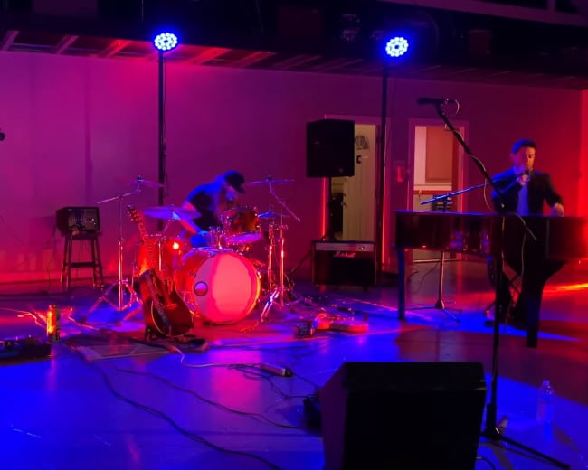 A band performs on stage with a bass guitar, drums, and piano, surrounded by colorful lighting and audio equipment.