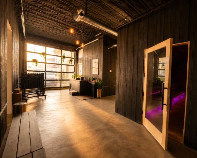 The image shows a modern interior space with wooden walls, concrete flooring, and large windows, featuring a reception area and a doorway leading to another room.