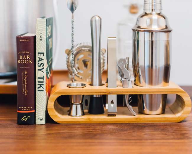 A wooden bar tool holder displays a cocktail shaker, strainer, and various mixing utensils alongside three cocktail recipe books.