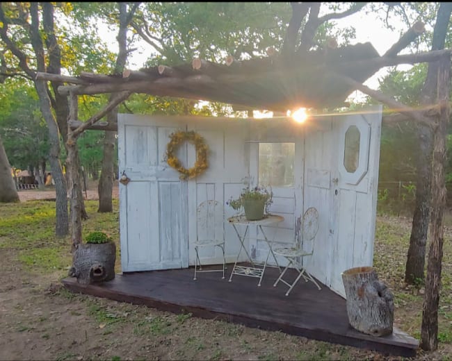 A rustic open structure with white wooden walls, a small table and chairs, and a wreath, is set in a wooded area with the sun rising in the background.