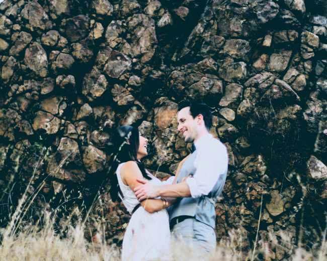 A couple embraces in a grassy area in front of a large rocky outcrop.