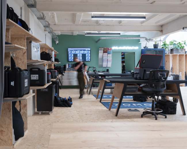 The image shows a spacious workspace with wooden shelving holding various gear, a large screen on the wall, and a desk area with a chair in a modern, industrial-style setting.