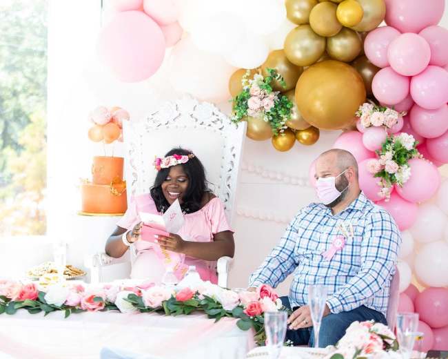 A woman in a pink dress reads from a booklet while sitting on an ornate chair next to a man in a plaid shirt during a festive gathering adorned with balloons and floral decorations.