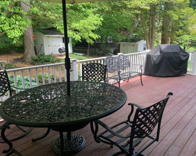 A patio area features a round metal table with chairs, an umbrella, and a grill, surrounded by lush greenery.