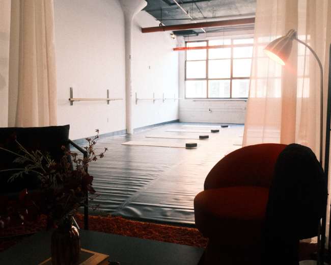 The image shows a spacious, softly lit studio with a dance floor, surrounded by sheer curtains and a seating area featuring a red chair and a table with a small floral arrangement.