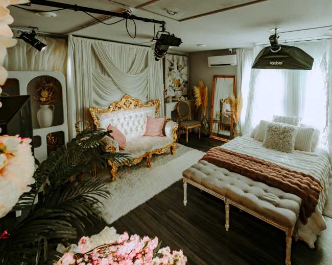 The image shows a decorated bedroom featuring a gold-accented sofa, a bed with a textured blanket, and soft, natural lighting from the windows.