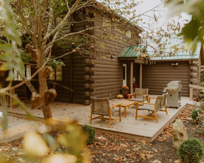 A wooden cabin features a patio with seating and a grill, surrounded by autumn leaves.
