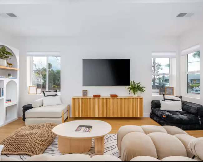 The image shows a modern, minimalist living room with a light color palette, featuring a large television mounted on the wall, a round coffee table, and various furniture pieces arranged around the space.